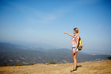 Young woman wanderer points finger on copy space while standing on a mountain hill against amazing view, female traveler enjoying beautiful landscape while taking break during hiking in summer day
