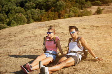 Tired man and woman travelers enjoying beautiful landscape while sitting on a hill in sunny summer day, young two wanderers relaxing outdoors after hiking in mountain during their vacation overseas