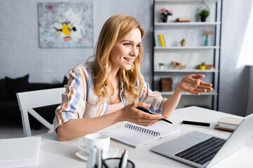 Selective focus of attractive woman smiling and gesturing near laptop