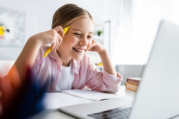 Selective focus of positive kid holding pen near notebook and looking at laptop on table