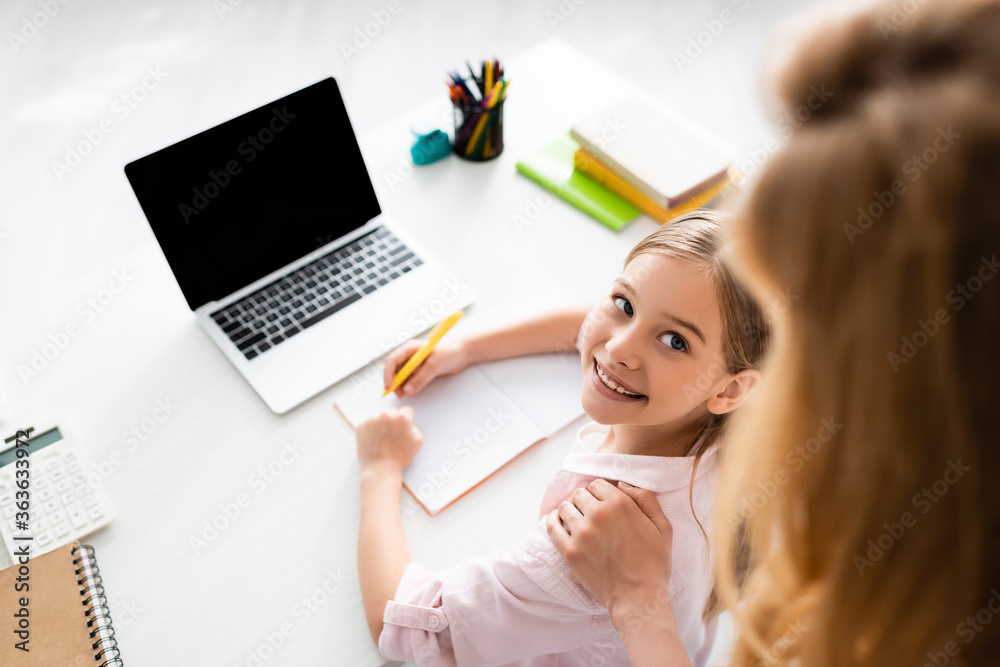 Wall mural overhead view of smiling kid looking at mother while writing on notebook near laptop on table