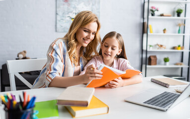 Fototapeta na wymiar Selective focus of smiling woman holding notebook while helping daughter during online education at home