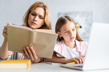 Selective focus of concentrated woman reading book near daughter using laptop at table