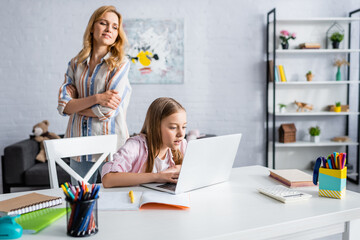 Selective focus of woman looking at daughter using laptop near stationery on table