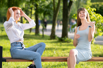 Two young girls friends sitting on a bench in summer park chatting happily having fun.