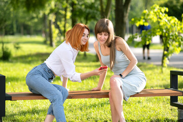 Fototapeta premium Two young girls friends sitting on a bench in summer park looking in red smartphone.