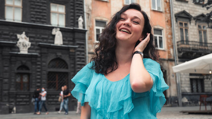 Fototapeta premium Young Caucasian woman with turquoise blouse curly hair standing im the street urban old city bacgkround. Portrait close up of young beautiful brunette woman.