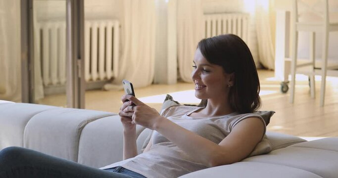 Smiling relaxed young woman holding smartphone texting sms message at home, happy girl using mobile apps for work education shopping chatting in social media online play game on phone sit on sofa