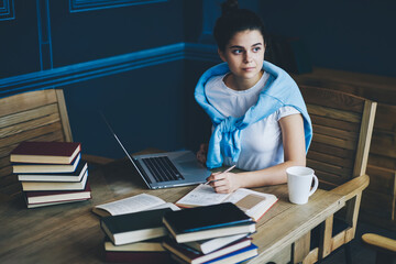 Pensive female student looking away while sitting at digital macbook computer and many literature books in stylish coworking space.Smart hipster girl preparing for training seminar in library