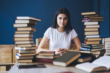 Portrait of skilled young woman holding modern smartphone in hands while looking at camera and sitting at table with many educational books in library.Clever student with cellular preparing for exams