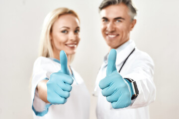 Two happy male and female doctors in medical uniform and blue gloves showing thumbs up, looking at camera and smiling, standing against grey background