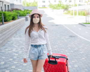 A beautiful young Caucasian woman in a medical mask walks down the street with a red suitcase. A girl with luggage travels alone during the height of the coronavirus epidemic. The empty city.