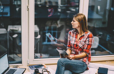 Pondering young woman in casual wear holding modern touch pad in hands while looking out of window sitting on windowsill and thinking on ideas for internet publication using 4G internet connection