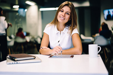 Portrait of cheerful young female student of art academie using modern technology for creating drawings,smiling hipster girl using stylus for writing on digital tablet sitting at table in library