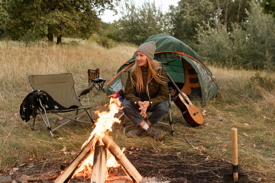 The blond girl in the autumn outfit sits near the firebond in the chair at the camp enjoying the atmoshere