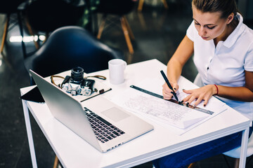 Professional female architect drafting blueprint accountings using ruler at desktop with netbook,...