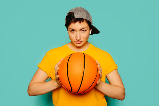 Portrait of defiant girl in sporty outfit over blue background holding a basketball ball.