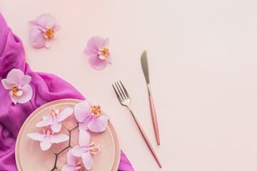 Flower and table settings overhead composition on light pink background. Pink ceramic plates, cutlery and perple orchid flowers. Holiday modern table decoration. Romantic date.