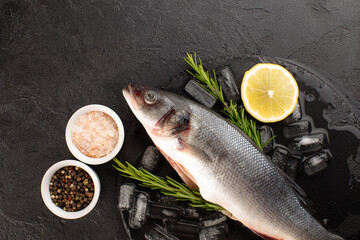 Fresh sea bass with ice cubes,rosemary,lemon,sea salt and dried pepper on black background.Top view.