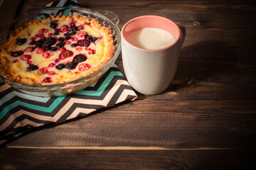 Baked round homemade cheesecake with berries is on a striped napkin. Pie and a mug of milk on an old dark wood table. Flatley with copyspace.