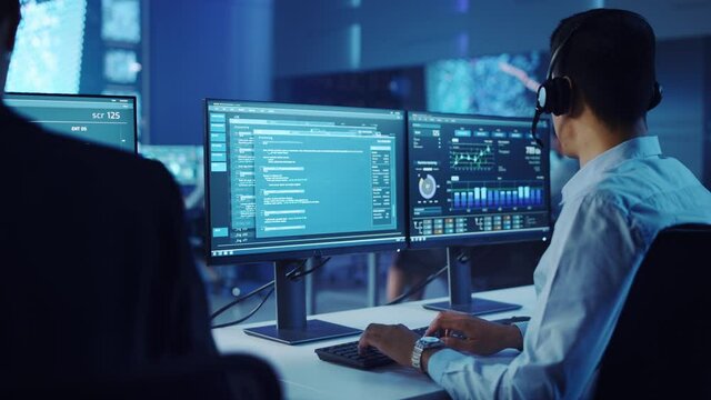 Confident Male Data Scientist Works on Personal Computer Wearing a Headset in Big Infrastructure Control and Monitoring Room. Young Engineer in a Call Center Office Room with Colleagues.