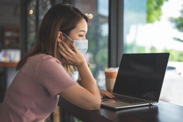 Asian woman wearing protective mask working on laptop in coffee shop while social distancing