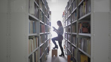 Young student girl taking book from shelf in university library.