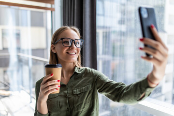Portrait of woman in formal wear standing holding takeaway coffee in hand and taking selfie on mobile phone in office