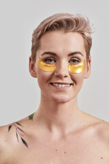 Portrait of half naked tattooed woman with short hair smiling, caring for her face, wearing under eye patches for dark circles isolated over light background