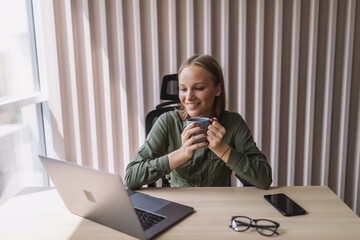 Young pretty woman drinking coffee at work place in front of laptop in office