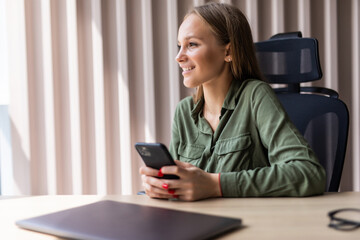 Young smiling business woman using smartphone near computer in office