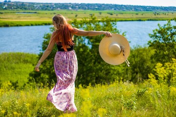 Cheerful girl with a hat in the meadow near the river. Countruside view.