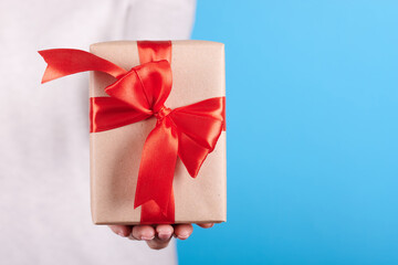 Present box with red ribbon in hand on blue background. Close up photo.