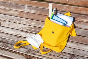 Yellow backpack with school supplies on a bench outdoors.Medical mask, virus protection. New school year, back to school