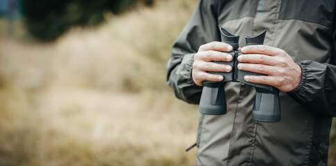 Panorama close up of a man dressed in a hunter jacket holds binoculars in his hand.