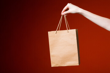 Female hand on a red background holds a paper gift bag. Online delivery and gift concept.