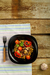 On a wooden table is a black dish masha with vegetables and mushrooms on a napkin and a fork. Copy space.