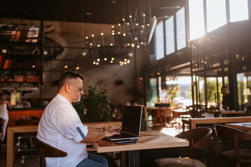 Side view of a caucasian middle-aged businessman with glasses and a white shirt while sitting in a cafe and working on a laptop.