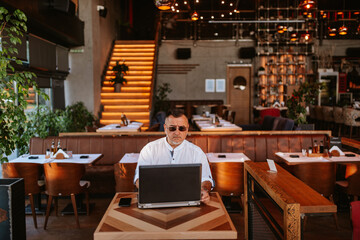 Portrait of a caucasian middle-aged man with glasses in a white shirt sitting in a cafe and working on a laptop