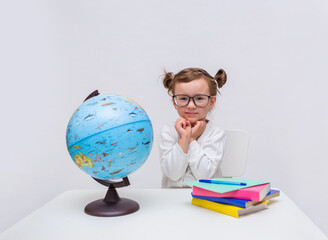 Fototapeta premium A cute little pupil sits at a table with textbooks and a globe on a white isolated background and looks at the camera