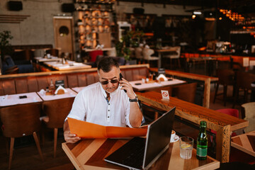 A middle-aged caucasian businessman with glasses in a white shirt is sitting in a cafe, talking on the phone and reading business documents