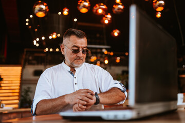 A middle-aged caucasian man with glasses in a white shirt sitting in a cafe looking at his watch while a laptop is on the table