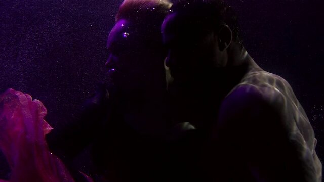 Close-up portrait of a tender couple in love who are under water and hugging, a buxom girl in a dress and a dark-skinned man. They are on a dark background with bubbles.