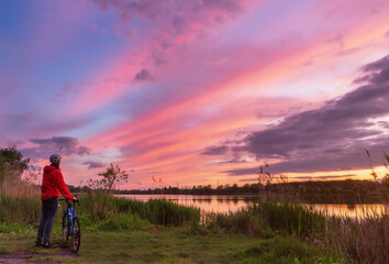 Fototapeta premium Cyclist on the coast of the river at sunset in summer. Landscape with man on gravel bike and colorful sky with clouds. Sport and travel concept.