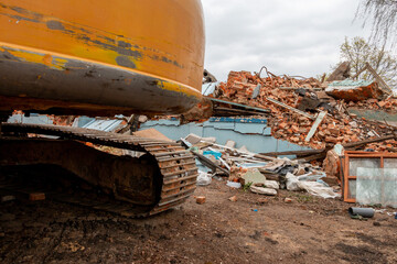 Fototapeta premium Excavator caterpillar on the background of a destroyed, brick, residential building. Demolition of an old house with an excavator.