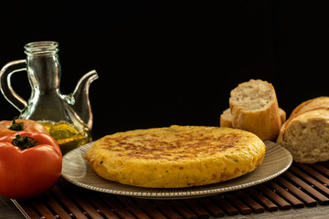 Spanish tortilla, with a glass jar of olive oil and some tomatoes and bread on a dark background