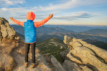 Young child boy hiker standing with raised hands in mountains enjoying view of amazing mountain...