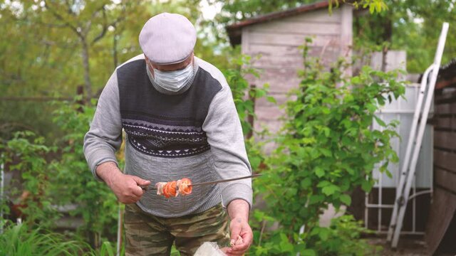 Mature man in medical mask impales raw shashlik on skewer. Older male holds skewer with barbecue. Concept of picnic outdoor.