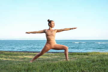 Young beautiful woman practices yoga on the grass by the ocean. Sporty yogi girl doing yoga doing war pose (virabhadrasana).