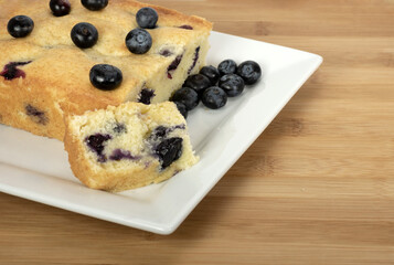 Homemade cream cheese blueberry bread and several loose blueberries are on a square white plate on a brown wooden board.
